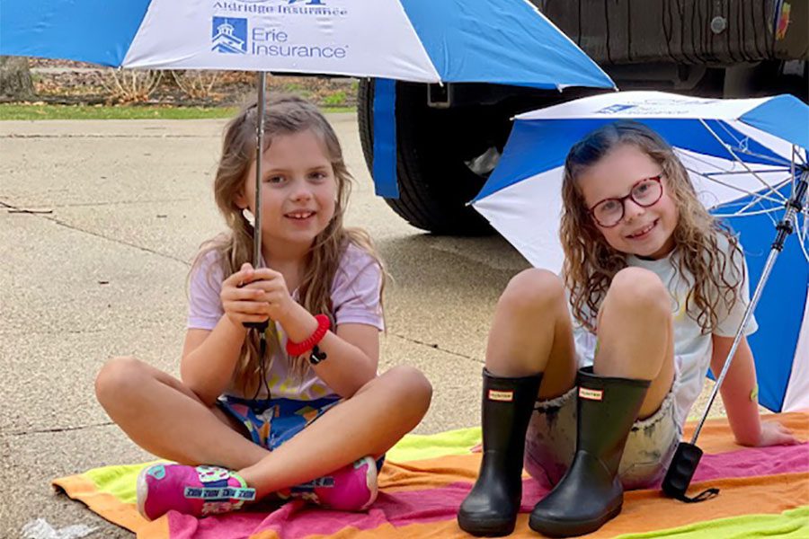 Umbrella Insurance - Portrait of Two Cheerful Little Girls Sitting on the Concrete Parking Lot on a Blanket While Holding Up Erie and Aldridge Insurance Umbrellas
